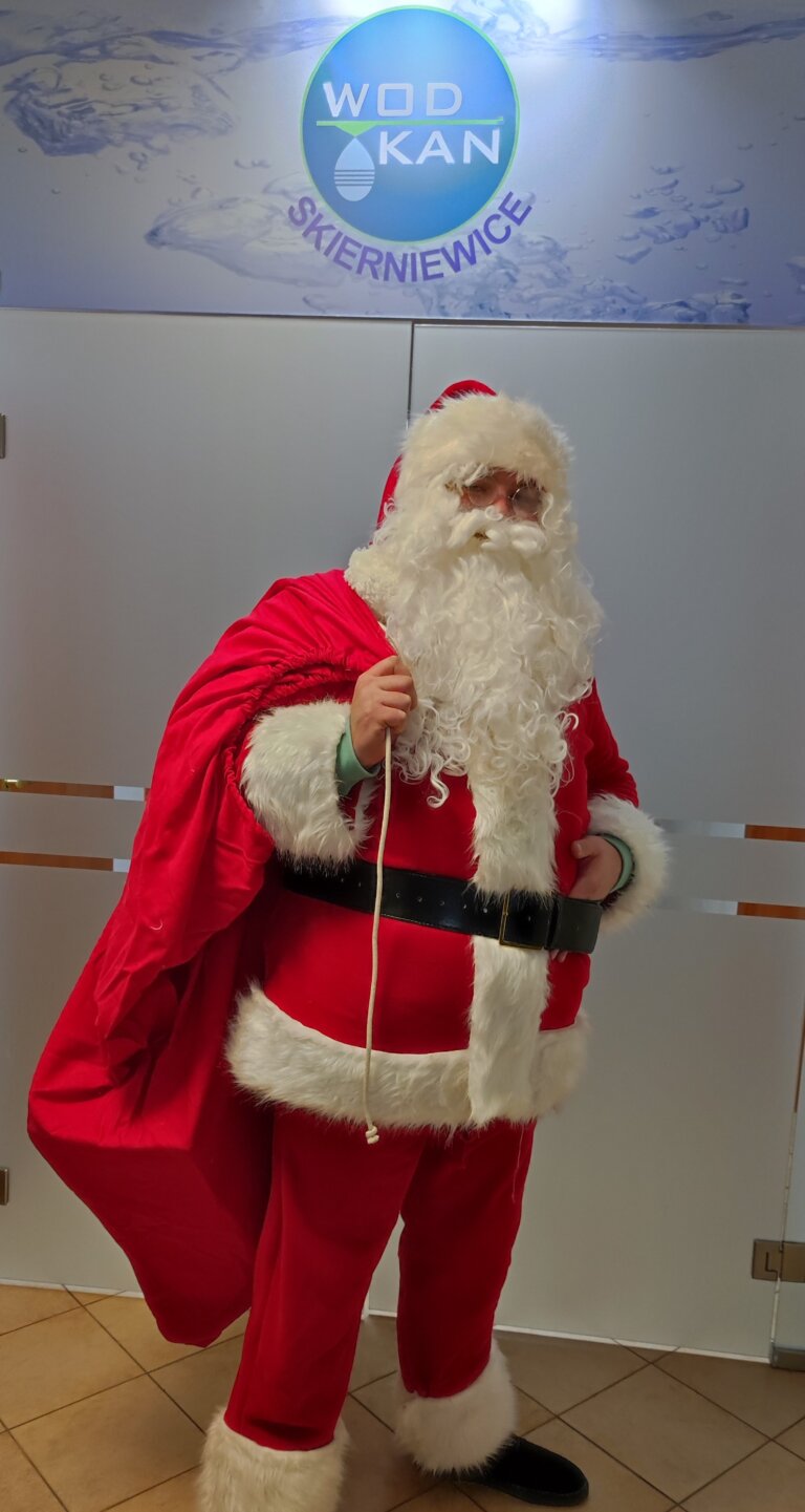 Na zdjęciu Mikołaj w czerwonym stroju, biała broda i czapka , na środku czarny pas przepasający brzuch. W ręce Mikołaj trzyma duży czerwony wór przerzucony przez ramię. Mikołaj pozuje na tle zamkniętych szklanych drzwi na górze logo i napis WOD-KAN