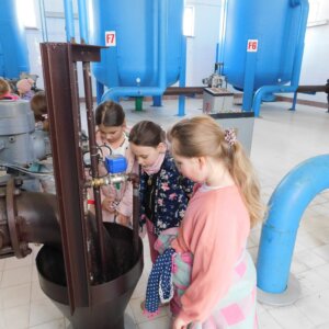 Trzy dziewczynki zaglądające do przepustników - specjalnych urządzeń w kształcie leja gdzie zbierają sie kroplu wody. W tle za dziewczynkami znajdują się niebieskie filtry wody z oznaczeniami f7,F6 znajdujących się w