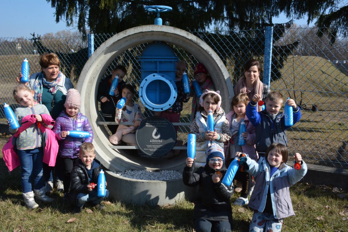 zdjęcie przedszkolaków z grupy "Kasztanki z Przedszkola nr 8 pozują na tle pomnika Iskierki z niebieskimi bidonami