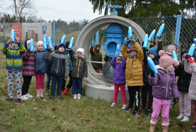 na zdjęciu grupa przedszkolaków pozująca z niebieskimi bidonami przy pomniku na Stacji Uzdatniania Wody przy ul. Waryńskiego w Skierniewicach