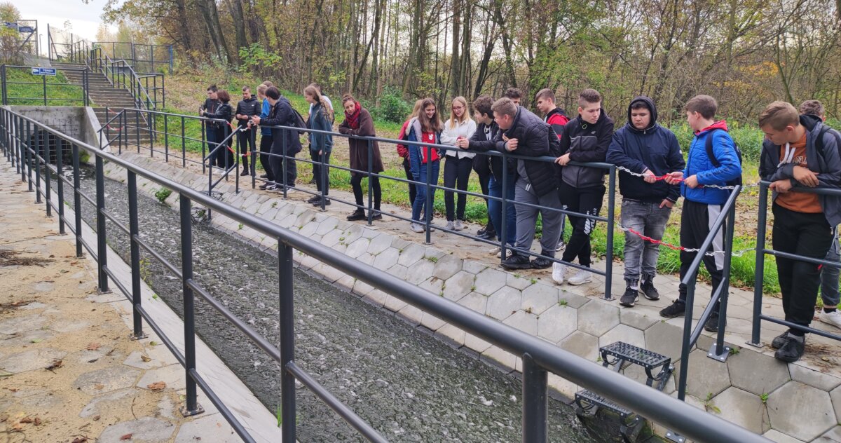 Na zdjęciu grupa młodzieży stojąca przy mostku i podziwiająca kanał którym wypływa woda